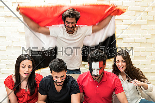 group of young people sitting on a sofa cheering for a football match in front of white table holding egyptian flags and one of them has egyptian flag paint on his face