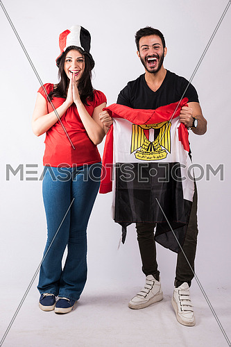 young couple standing and cheering holding big egyptian flag, while the female wearing egyptian flag hat on white background