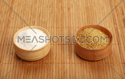 Choice, selection of two sugars in small full wooden bowls on bamboo mat background, brown cane and white sugar