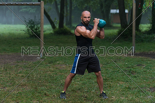 Strong And Muscular Young Fitness Man -  Holds Up A Green Kettlebell Outdoors  - Sports And Fitness - Concept Of Healthy Lifestyle - Fitness Male