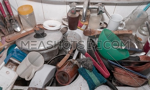 From above of heap of messy dishes stacked in metal sink in dirty rustic kitchen