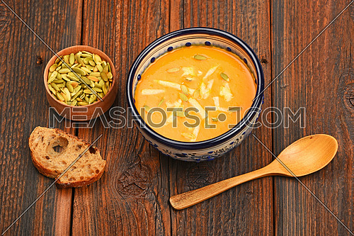 Ceramic bowl of pumpkin cream soup, spoon, slice of bread and seeds on dark wooden table background, high angle view