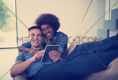Young multiethnic couple relaxing at luxurious home with tablet computers reading in the living room on the sofa couch.