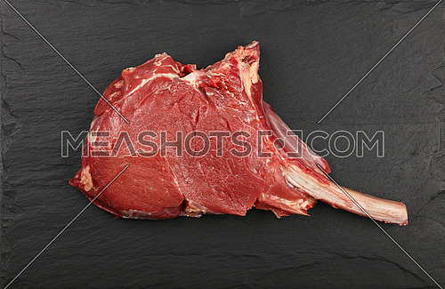 Close up one raw tomahawk ribeye beef steak with rib bone on black slate board background, elevated top view, directly above