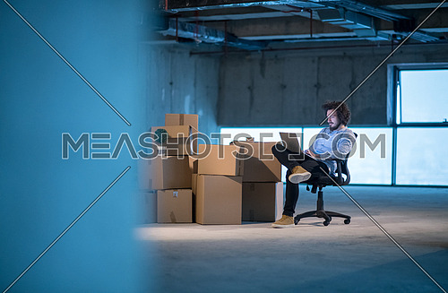 young male architect on construction site checking documents and business workflow using laptop computer with cardboard boxes around him in new startup office