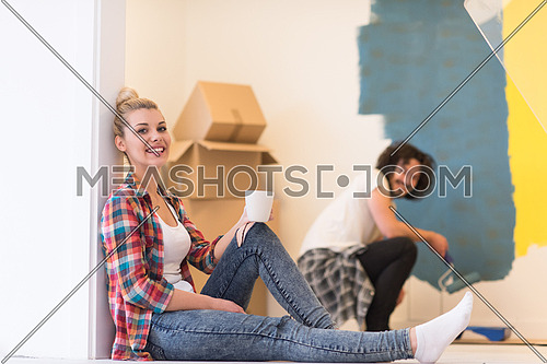 Happy couple doing home renovations, the man is painting the room and the woman is relaxing on the floor and drinking coffee