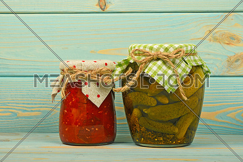 One glass jar of homemade eggplant pepper salad and pickled cucumbers with green checkered textile decoration over blue painted wooden surface