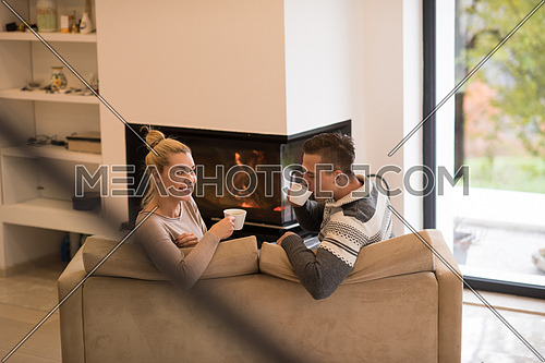 Young romantic couple sitting on sofa in front of fireplace at home, looking at each other, talking and drinking coffee at autumn day