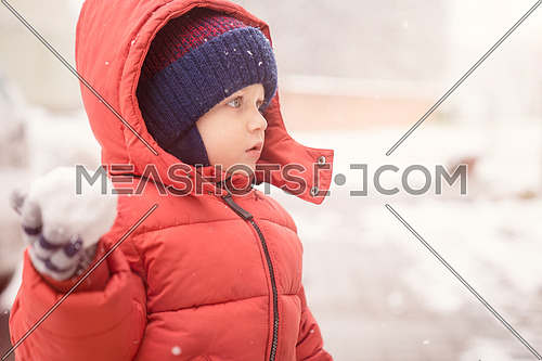 Infant boy while snowing looks towards the emptiness, holding in his hand a snowball, covered with red winter jacket and woolen hat, close-up.
