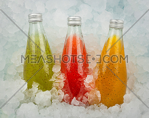 Close up three glass bottles of cold orange, red and green juice cocktail drinks with chia seeds on crushed ice at retail display, low angle side view