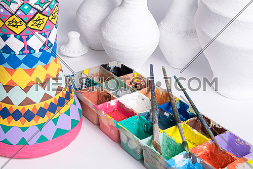 Part of a pottery painting art studio consists of color palette, brushes, and unfinished pottery craft vases on white background