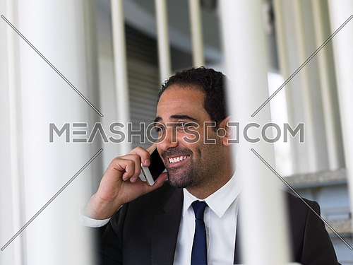 handsome young middle eastern business man working on laptop computer online while sitting on steps in front of modern corporate office building and speaking by phone
