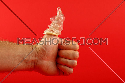 Close up man hand showing thumb up gesture with latex condom on finger over red background with copy space, low angle side view