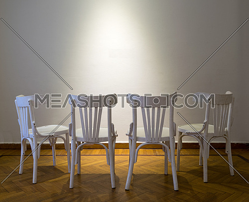 Four white wooden chairs facing spot lighted white wall on wooden parquet floor