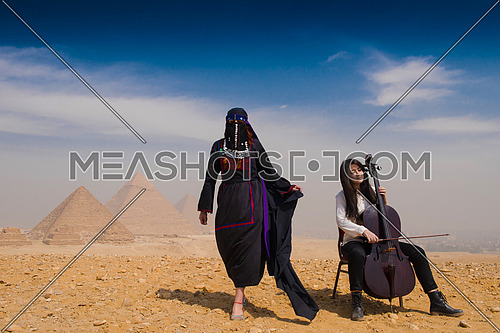 famale chello player and woman in traditional egyptian clothes in  ftont of pyramids