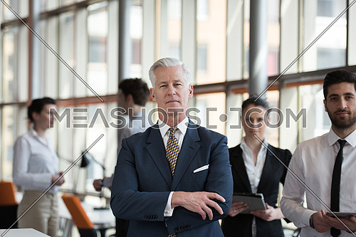 portrait of senior businessman as leader  at modern bright office interior, young  people group in background as team