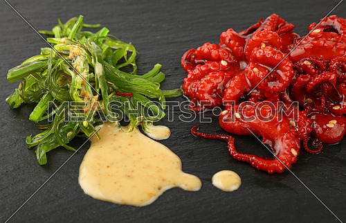 Seafood small whole red baby octopus salad with green seaweeds and peanut sauce on black slate board, close up, high angle view
