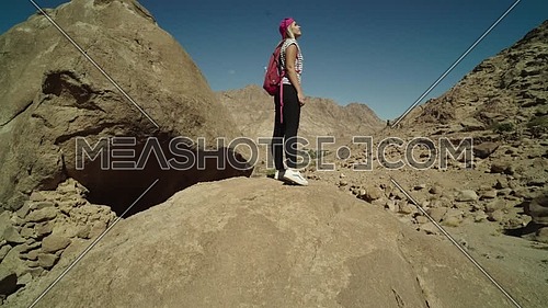 Track In shot for a female tourist wearing a pink cap and travel backpack standing upon a big rock to explore Sinai Mountain for wadi Freij at day.