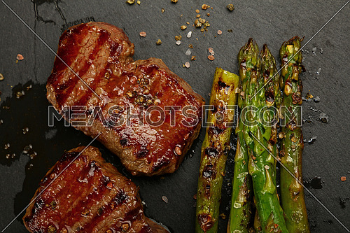 Close up one grilled roasted beef steak with green asparagus on black slate cutting board, elevated top view, directly above
