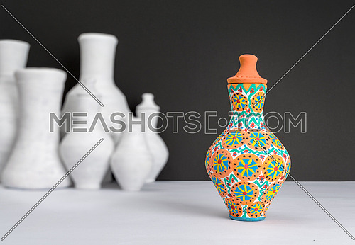 Still life of one decorated painted colorful pottery vase on background of blurred group of white vases, white table and black wall
