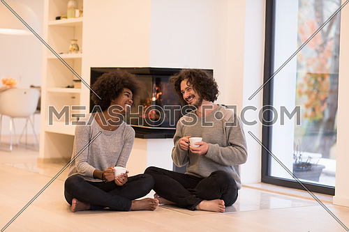 Young romantic multiethnic couple sitting on the floor in front of fireplace at home, looking at each other, talking and drinking coffee at autumn day