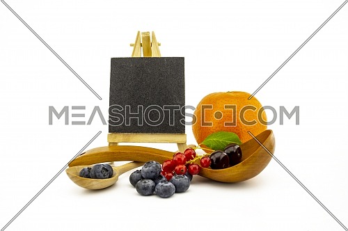 Fresh seasonal fruit still life with small chalkboard and assorted berries including blueberries, cherries and red currants on wooden spoons and orange on a white background