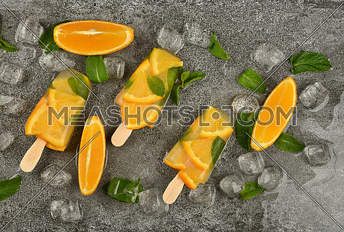Close up three fruit ice cream popsicles with fresh orange slices, green mint leaves and ice cubes on gray table surface, elevated top view, directly above