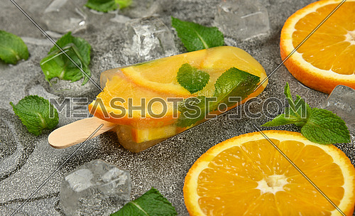 Close up one fruit ice cream popsicle with fresh orange slices, green mint leaves and ice cubes on gray table surface, high angle view