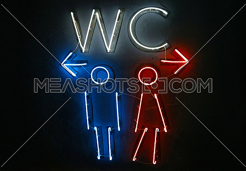 Close up glowing blue and red neon lights of WC toilet sign with directions to men and women, left and right arrows, over dark black wall background