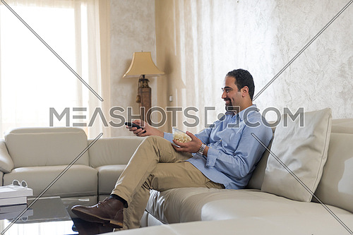 young middle eastern man enjoys with remote control and popcorn on the sofa