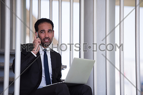 Young middle eastern business executive sitting on the stairs of a corporate office working with his laptop and talking on the phone