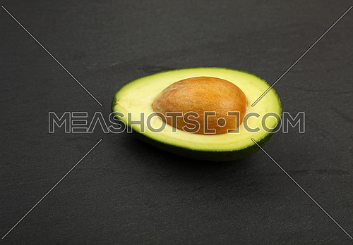 One fresh ripe green avocado half with pit stone on black slate board background, detail, close up, high angle view