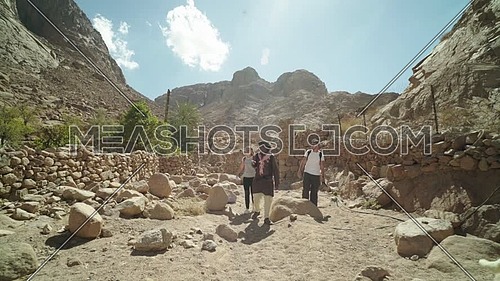Follow shot for group of tourists walking on big rocks between two fences of rocks with bedouin guide showing almond trees while explore Sinai Mountain for wadi Freij at day.
