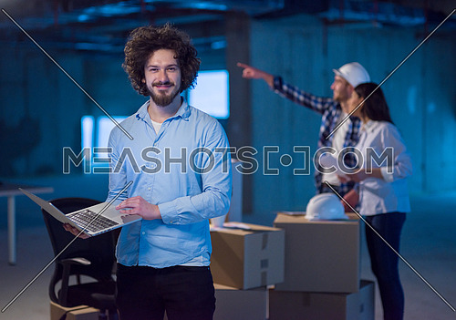 young male architect on construction site checking documents and business workflow using laptop computer with colleagues in the background in new startup office