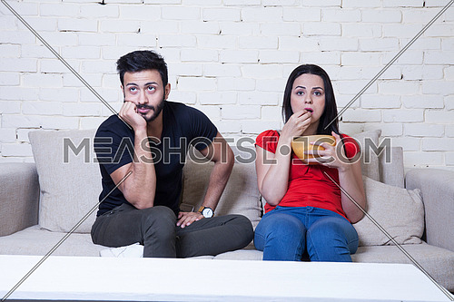 A young man and a young woman sitting on a cough eating popcorn