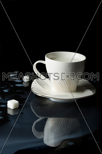 close up shot for a cup of milk put on black reflective surface and milk is poured around cup with two cube of sugar.
