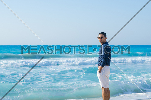 a young man wearing shorts and shirt standing looking towards the sea