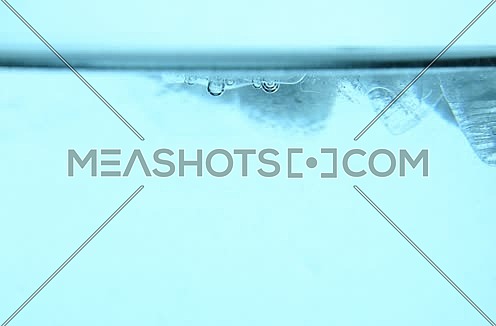 Clear ice cubes with bubbles of air float, spin, whirl and melt in blue water, close up, low angle side view, slow motion