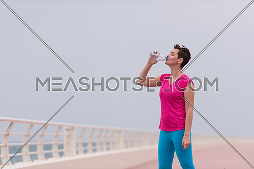 Fitness woman drinking water after running at the promenade by the sea. Thirsty sport runner resting taking a break with water bottle drink outside after training. Beautiful fit sporty girl.