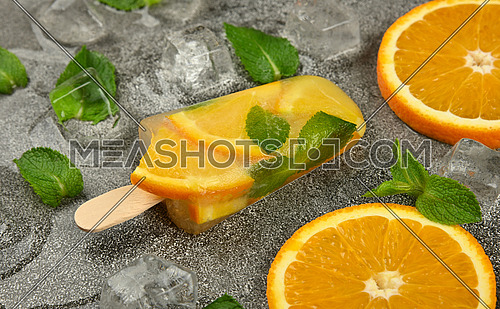 Close up one fruit ice cream popsicle with fresh orange slices, green mint leaves and ice cubes on gray table surface, elevated top view, directly above