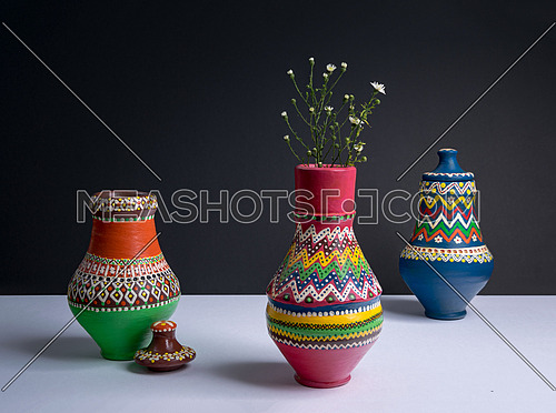 Studio shot of still life of three orange decorated pottery vases with small flowers in a background of white table and black wall with harsh shadow