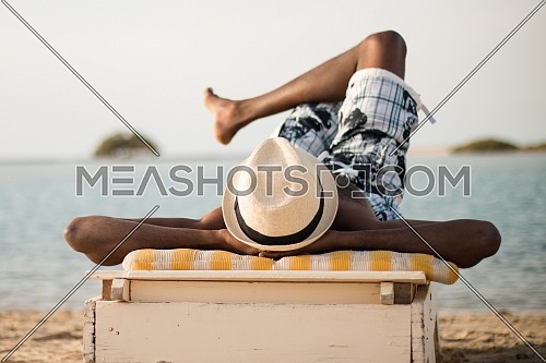 Long shot for a young man relaxing on Beach Chaise Lounge Chairs in front of the Red Sea wearing shorts and fedora hat at day