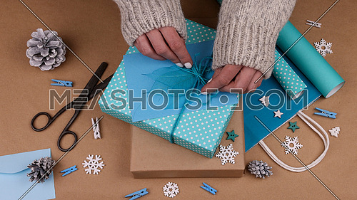 Close up woman hands packing and wrapping Christmas gift boxes with blue paper, adding envelope and skeleton leaves, high angle view