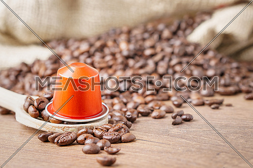 In the foreground a coffee capsule on wooden spoon and  roasted coffee beans with burlap sack on blur wooden background ,close up.