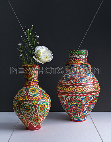 Still life composition of two colorful pottery vases, white flower, and small flowers on background of white table and dark wall