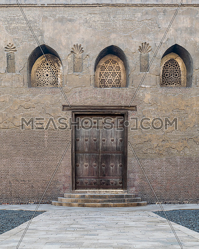 Aged wooden weathered door, perforated arched stucco window decorated with floral patterns, and three steps on stone bricks wall, External facade of Ibn Tulun Mosque, Cairo, Egypt