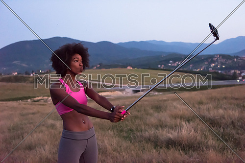 Healthy Looking Smiling Young African American Female in sports apparel photographing herself with a selfie stick beautiful summer evening in nature