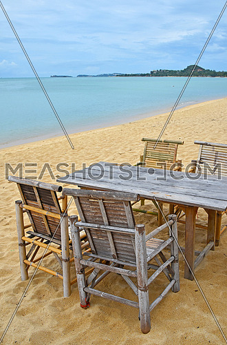 Wooden bamboo furniture, table and four chairs on sand beach with blue sea water and cloudy sky background