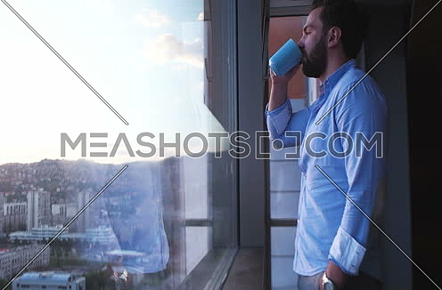 Man in shirt drinking coffee or tea by window admiring greath view on town during sunset