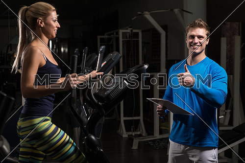 Young Woman Doing Aerobics Elliptical Walker In Gym And Personal Trainer Watching On Her